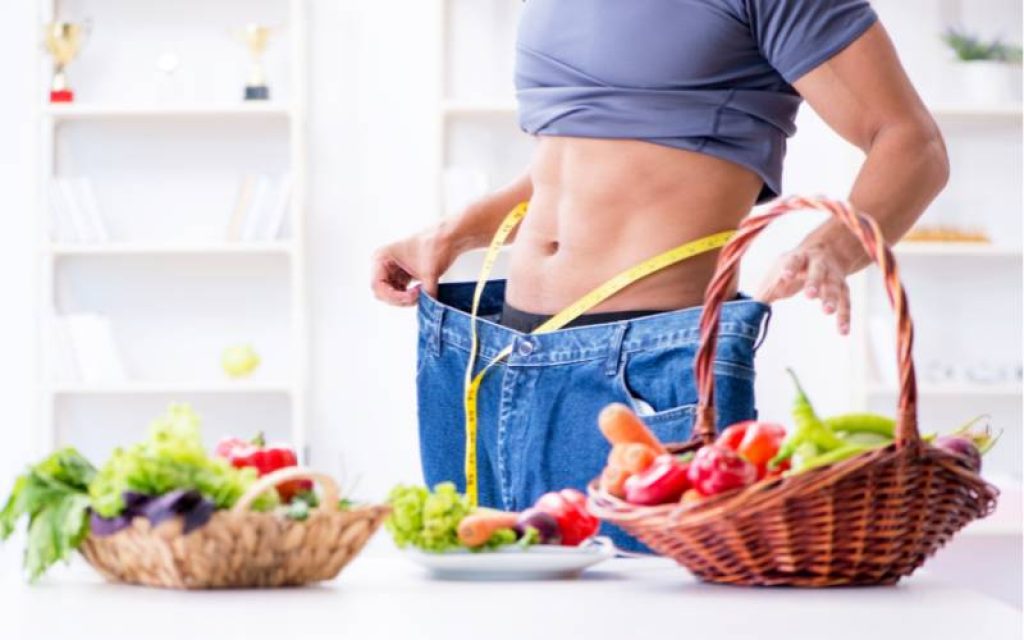 Pessoa com uma calça jeans larga e uma fita métrica ao redor da barriga próxima de pratos e cestas com frutas e verduras